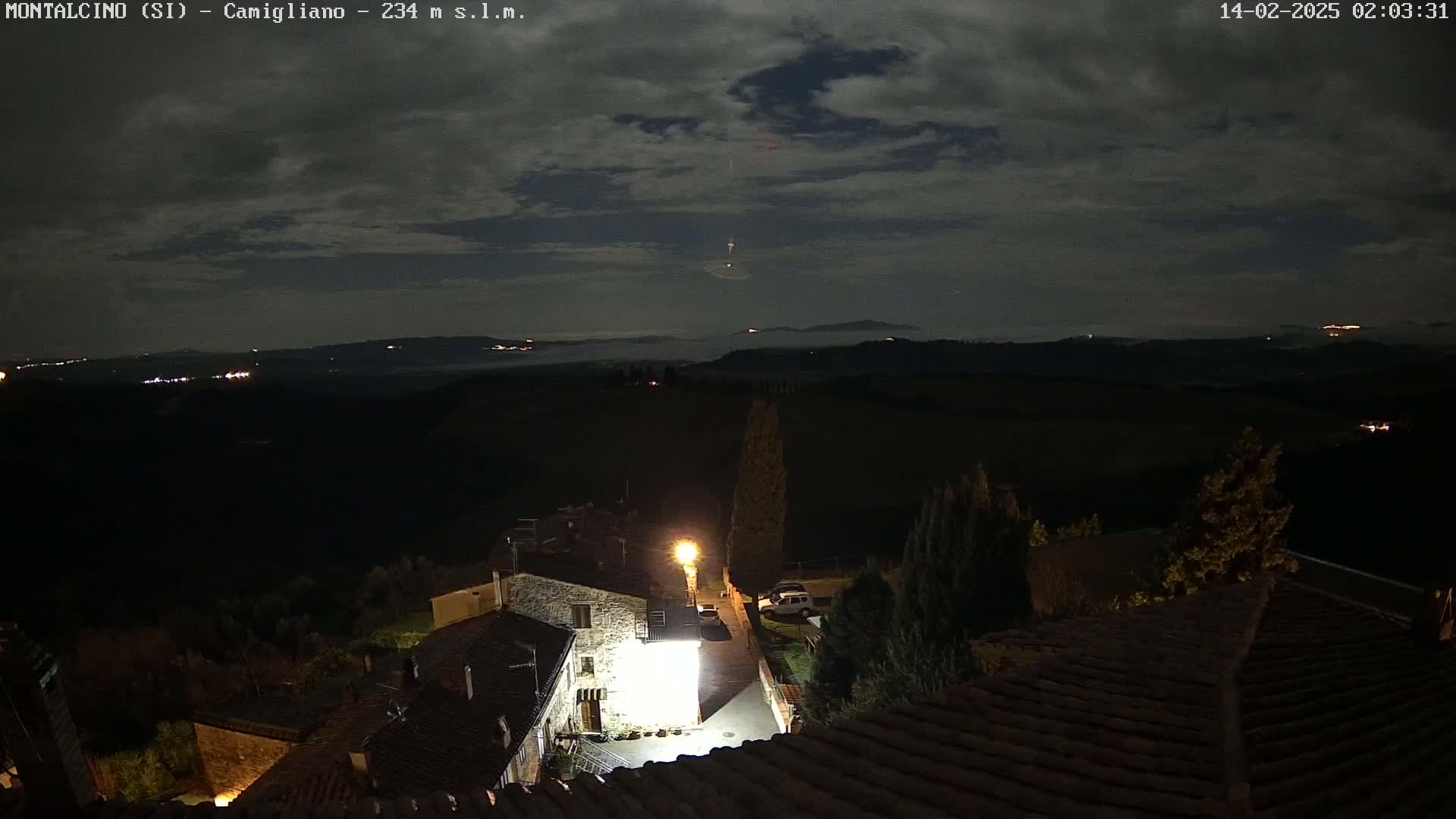 A nighttime view of a small village nestled in a hilly landscape under a partly cloudy sky, with a single light illuminating a courtyard.