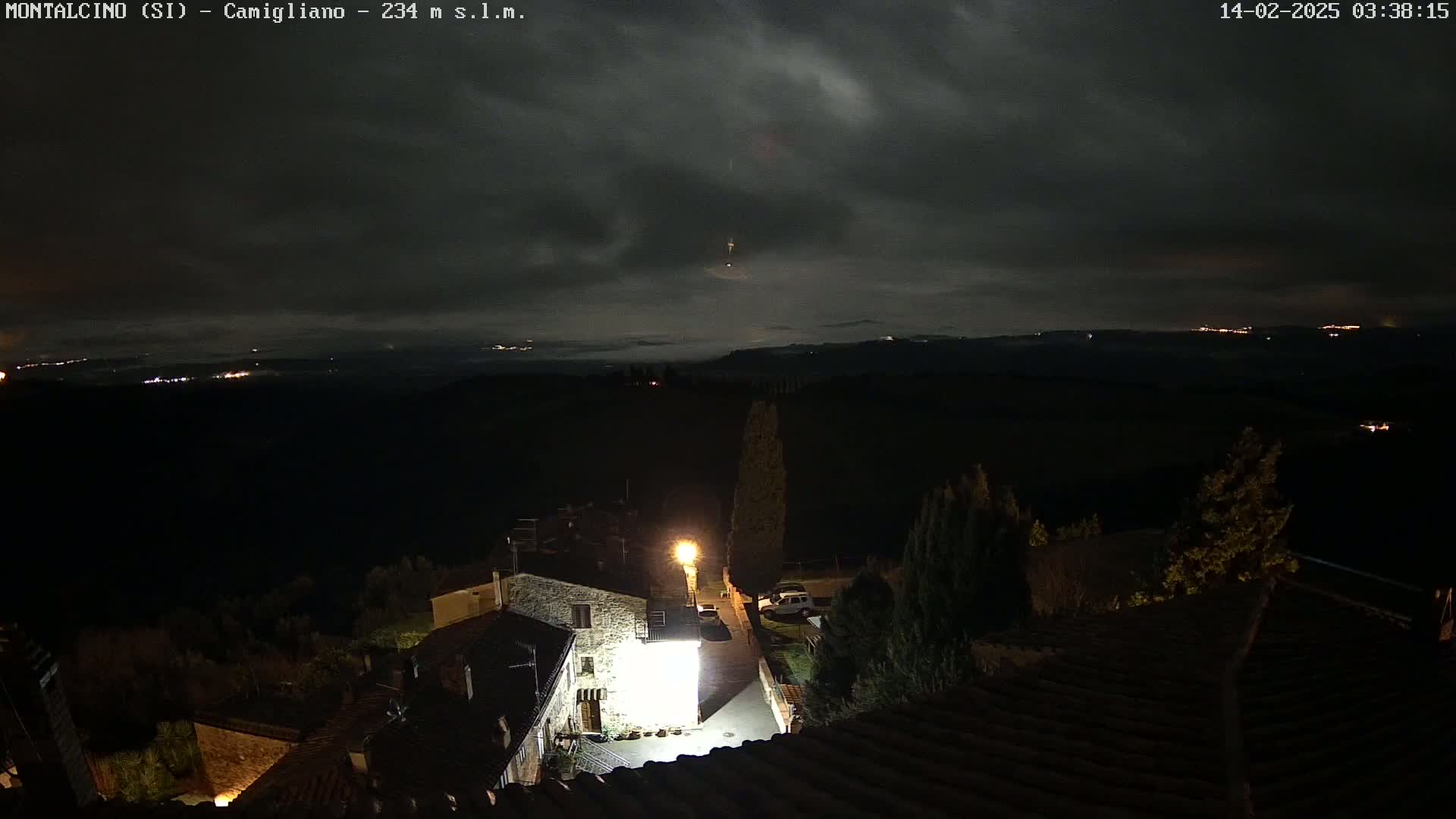 A nighttime view of a small village nestled in a hilly landscape under a cloudy sky, with distant lights visible on the horizon.