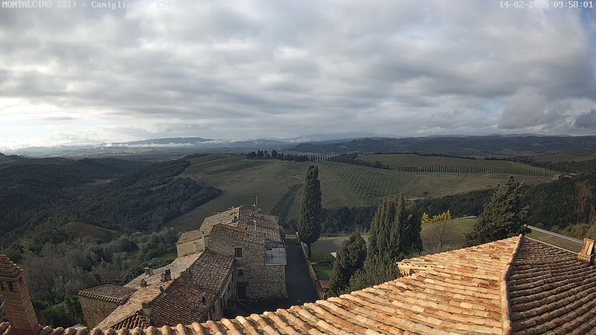 A view from a rooftop shows a cloudy sky over a hilly, tree-dotted landscape, with stone buildings and terracotta roofs in the foreground.