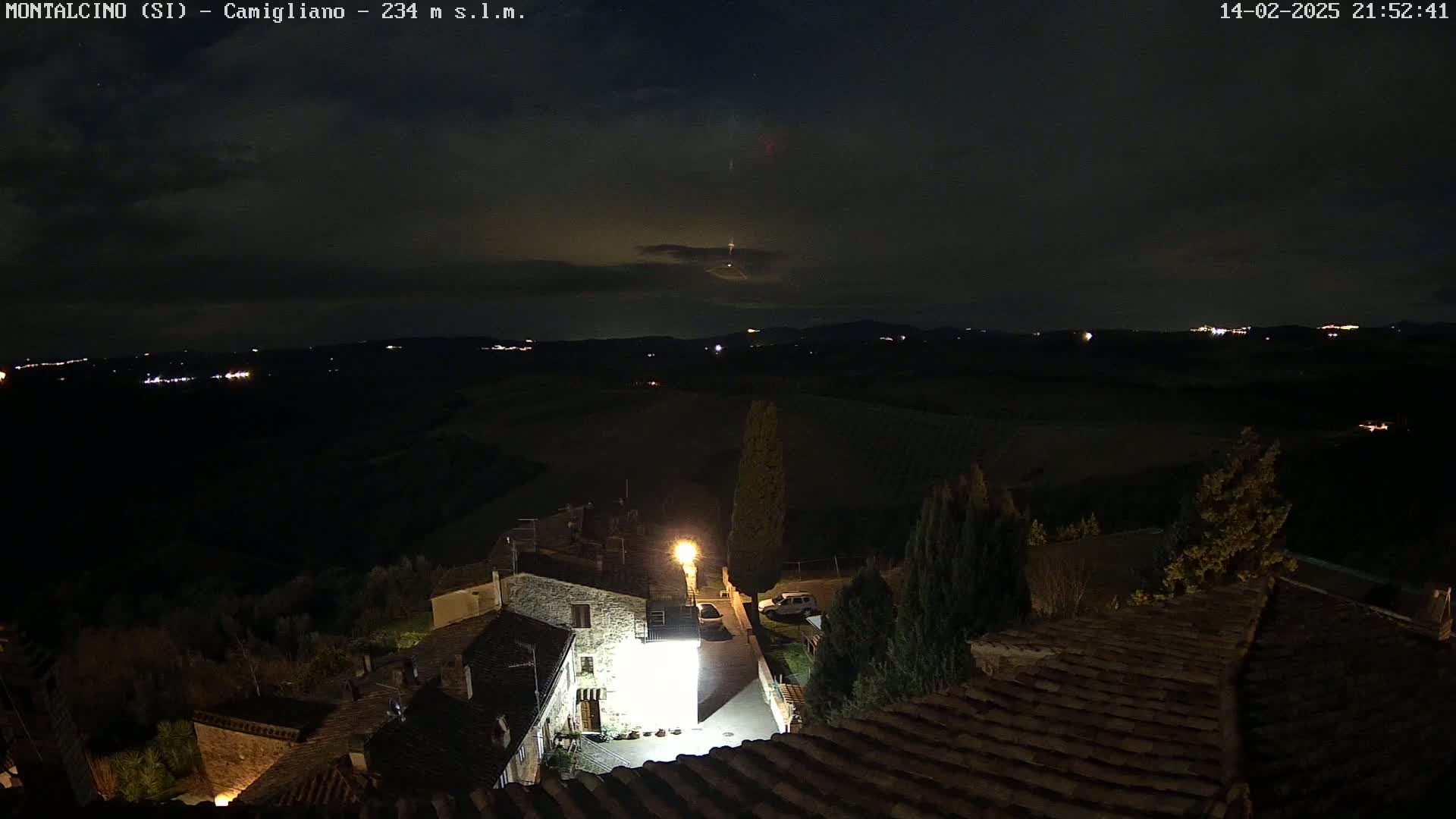 A nighttime view of a village illuminated by streetlights, with a dark, cloudy sky and distant lights visible on a hilly landscape.