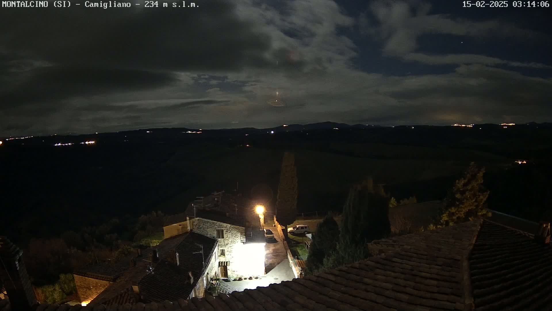 A nighttime view of a small village nestled in a hilly landscape under a partly cloudy sky.