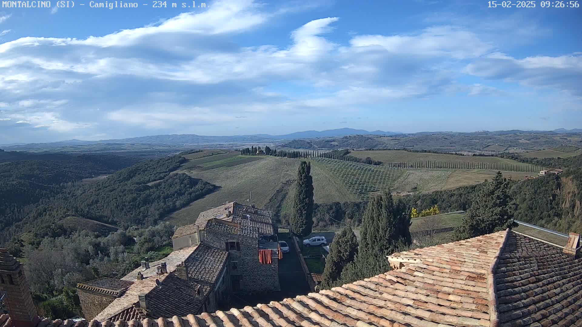 A view from a rooftop shows terracotta-tiled roofs of buildings overlooking a hilly landscape with rows of trees under a mostly clear, blue sky with scattered clouds.
