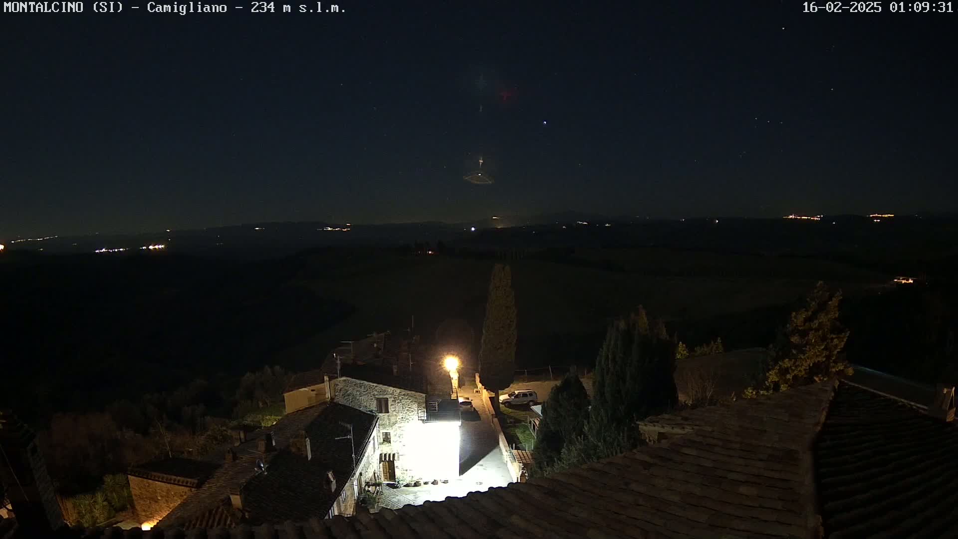 A nighttime view of a small village with a few lit buildings and streets, under a clear, starry sky overlooking a dark, distant landscape.