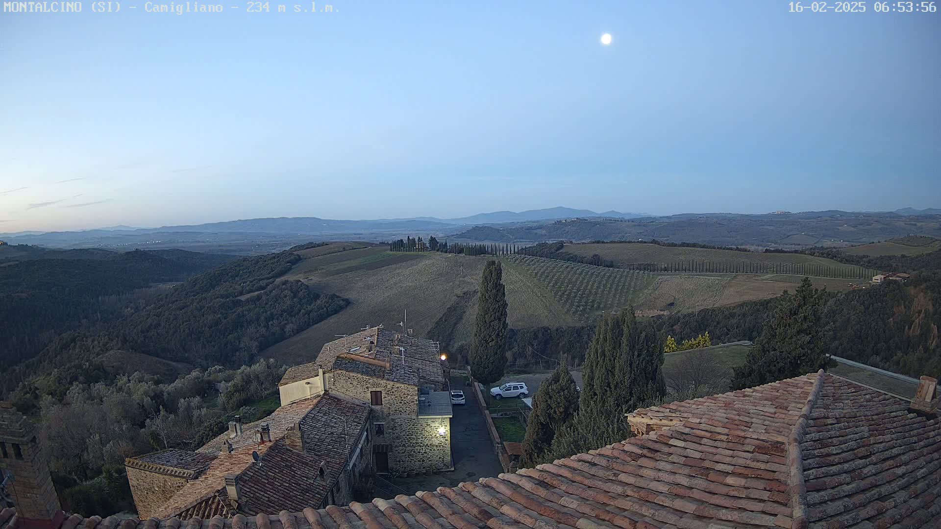 A moonlit, twilight view overlooks a hilly, Tuscan landscape of olive groves and stone buildings with terracotta roofs.