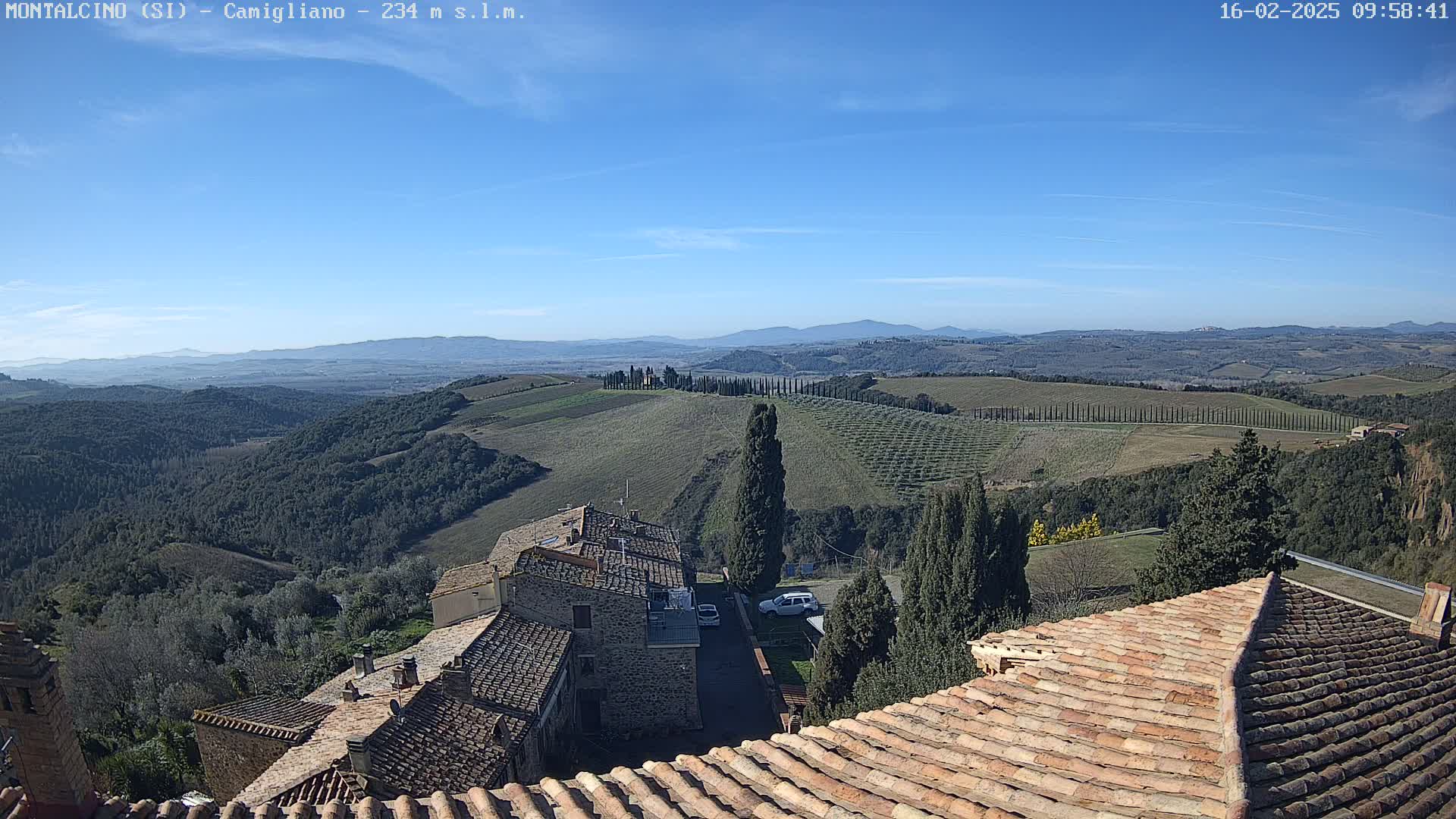 A clear, sunny day reveals a picturesque Tuscan landscape of rolling hills, vineyards, and cypress trees, viewed from atop a building with terracotta tile roofs.