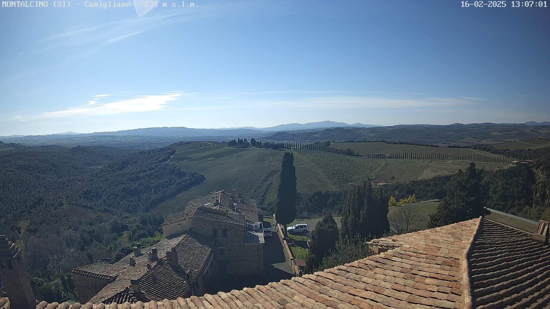 A sunny, clear day reveals a panoramic view of rolling hills, vineyards, and cypress trees, overlooking terracotta rooftops of buildings nestled in the Tuscan landscape.