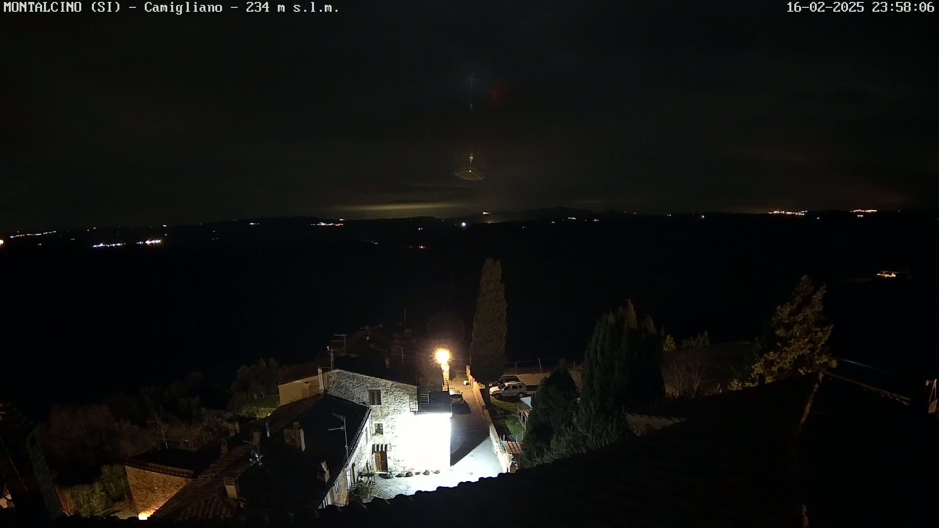 A nighttime view of a village illuminated by streetlights, overlooking a dark landscape with distant lights under a mostly clear sky.