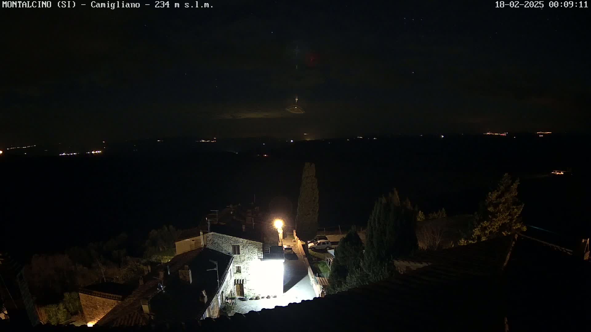 A nighttime view of a small village illuminated by a single bright light, with a dark, starry sky and distant lights visible in the background.