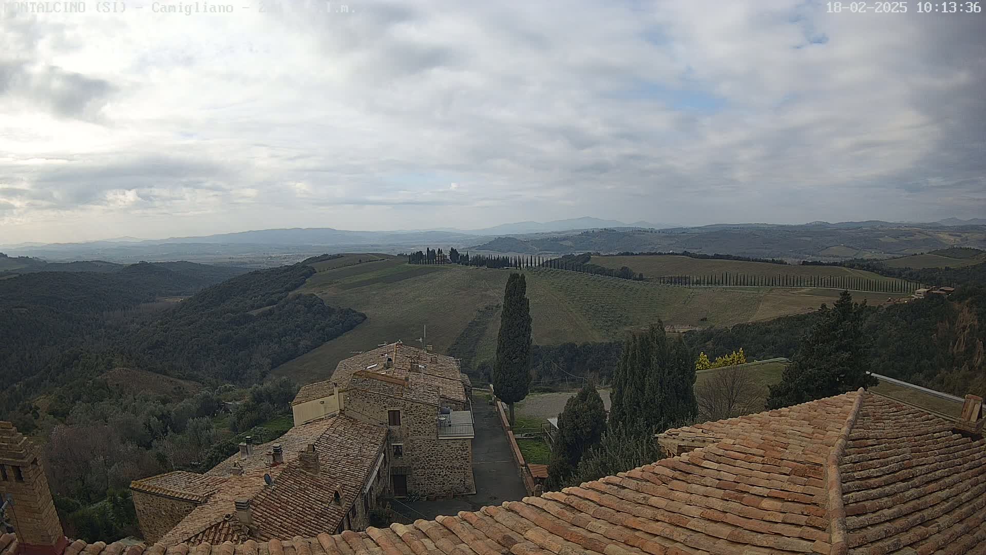 A view from a rooftop shows rolling hills, farmlands, and a line of cypress trees under a cloudy sky.