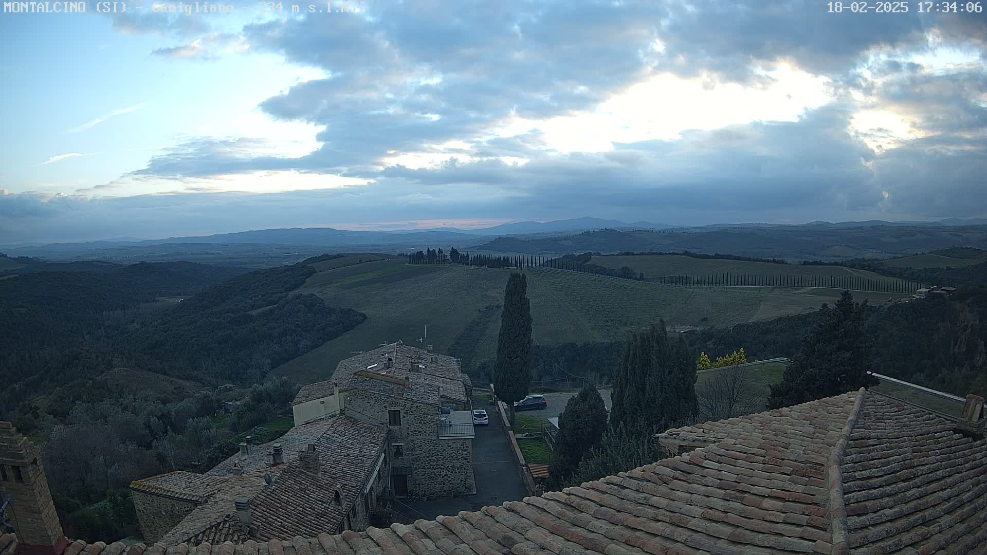 A view from a rooftop shows a cloudy twilight sky over rolling hills dotted with cypress trees and a small town with terracotta roofs.
