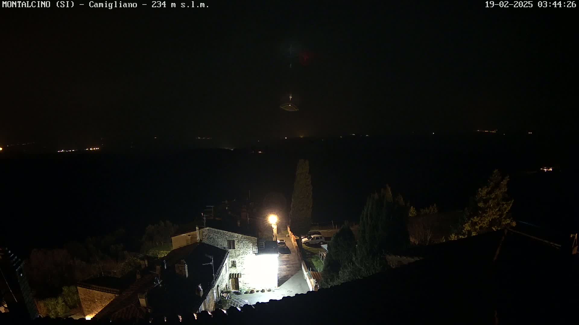 A nighttime view of a village with stone buildings illuminated by a single bright light, overlooking a dark landscape under a clear sky.