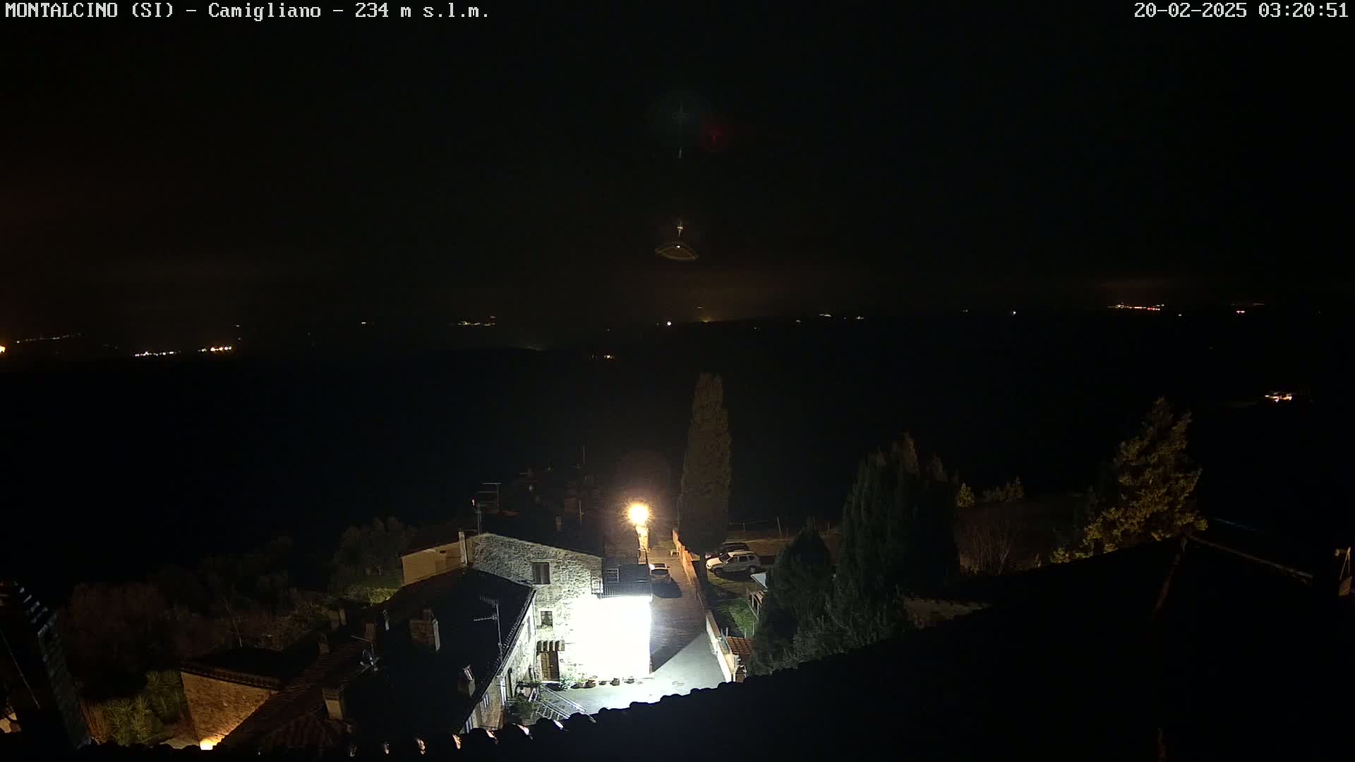 A nighttime view of a small village illuminated by a single bright light, overlooking a dark landscape with distant lights under a clear, dark sky.