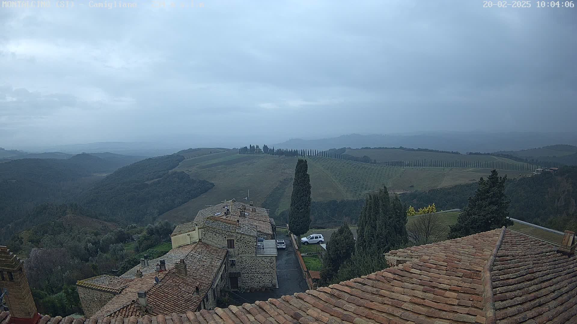 An overcast view from a rooftop shows a stone village nestled in rolling hills dotted with cypress trees under a gray sky.