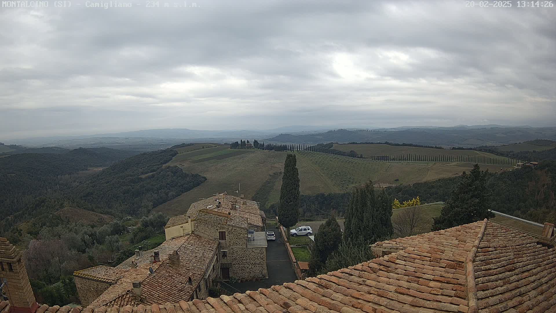 Under a cloudy sky, a view overlooking terracotta-roofed buildings and rolling hills dotted with cypress trees extends to a distant, hazy valley.