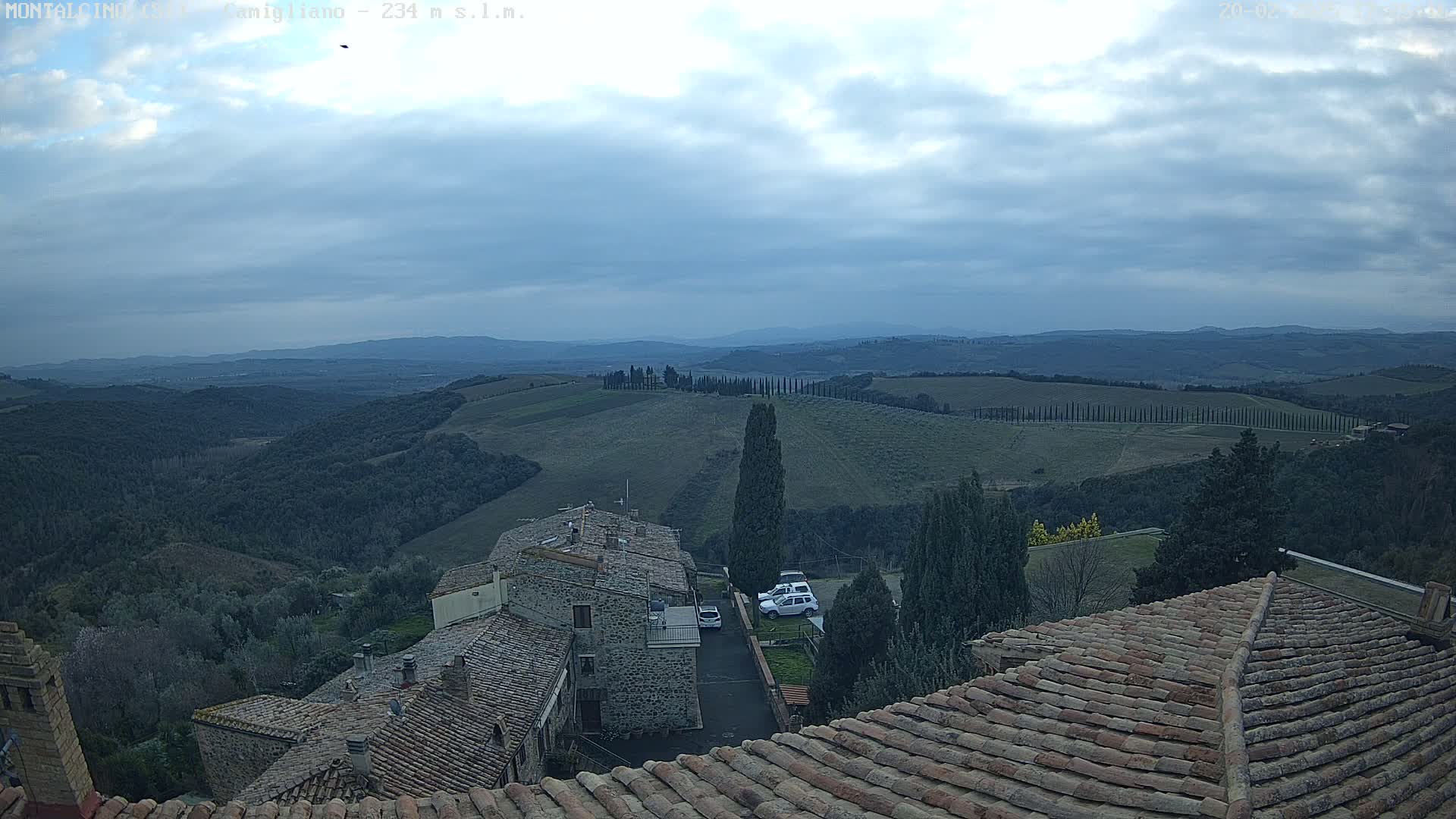 Under a mostly cloudy sky, a view from a rooftop shows tiled roofs of buildings overlooking a hilly landscape with rows of trees and distant mountains.