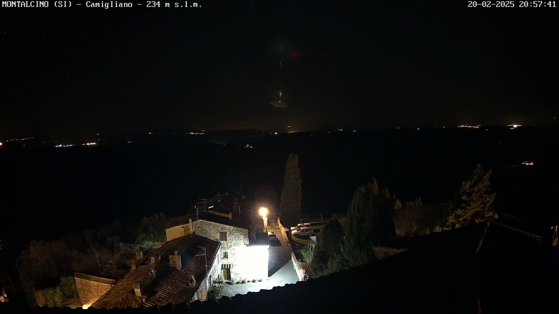 A nighttime, high-angle view of a small village illuminated by lights, with a dark, starlit sky and distant lights visible in the background.