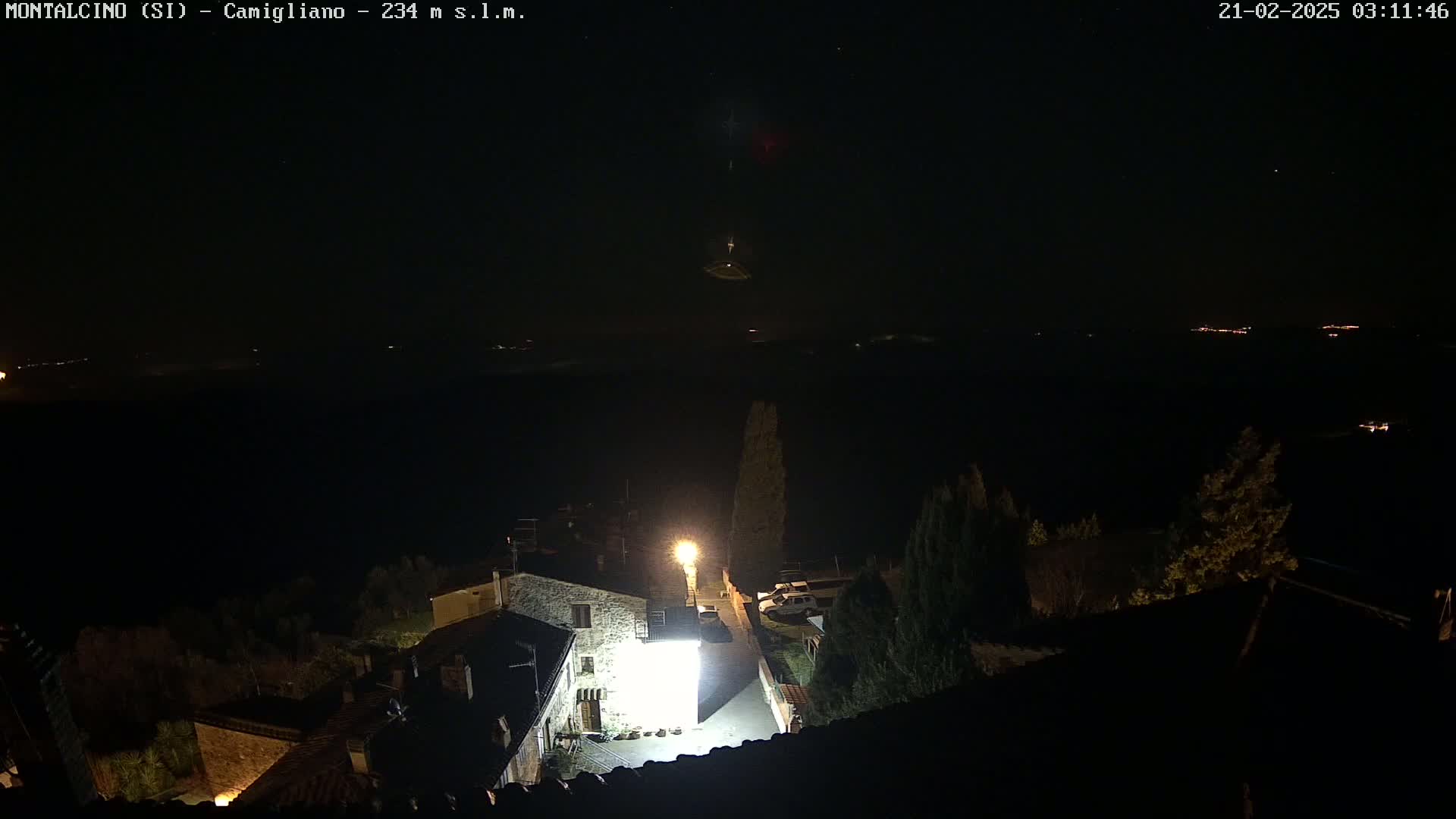A nighttime view of a small village illuminated by a single bright light, overlooking a dark landscape under a clear, starlit sky.