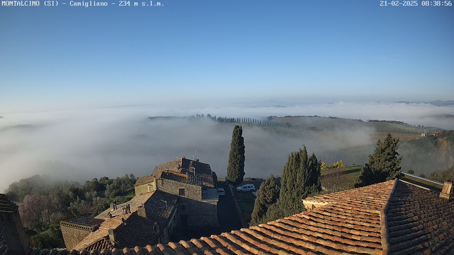 A fog-shrouded valley, visible from atop a village of stone buildings with terracotta roofs, under a clear blue sky.