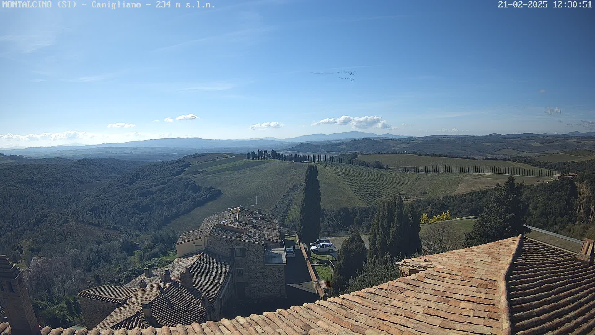 A view from a terracotta roof shows a sunny, hilly landscape with rows of cypress trees, stone buildings, and distant mountains under a mostly clear sky.