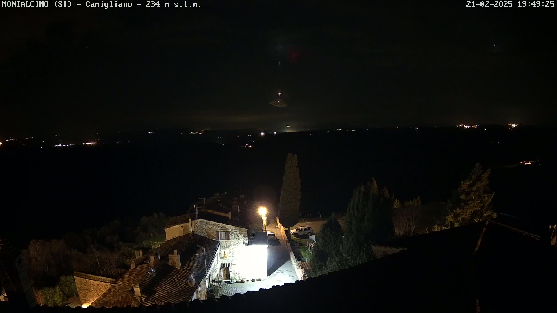 A nighttime view of a small village with stone buildings illuminated by streetlights, overlooking a dark, distant landscape under a mostly clear sky.