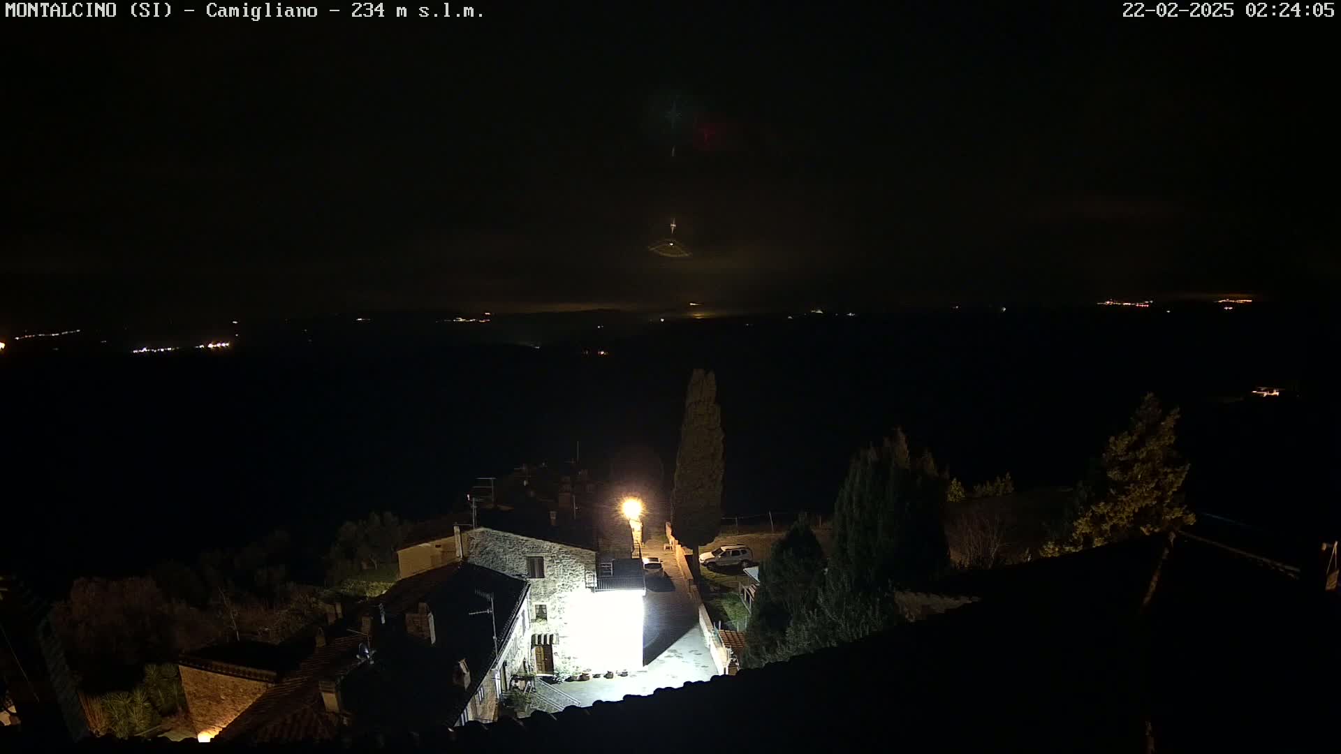 A nighttime view of a village illuminated by a bright light, with distant lights and what appears to be a faint fog or haze in the distance under a dark, clear sky.