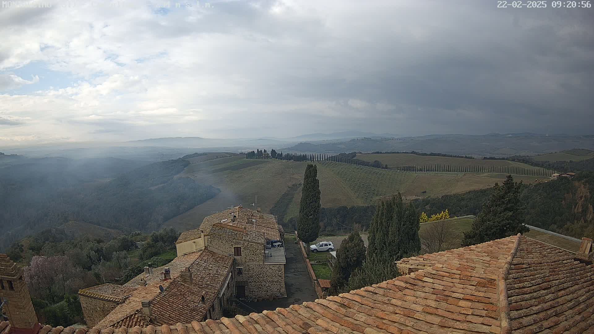 Overcast skies reveal a view of rolling hills and stone buildings with terracotta roofs, all under a mostly cloudy sky.