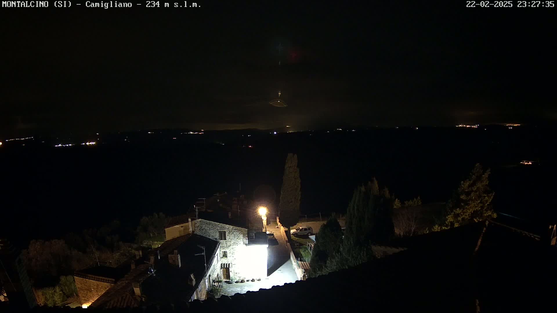 A nighttime, high-angle view shows a village with a few lit buildings and trees, overlooking a dark, distant landscape with faint lights.