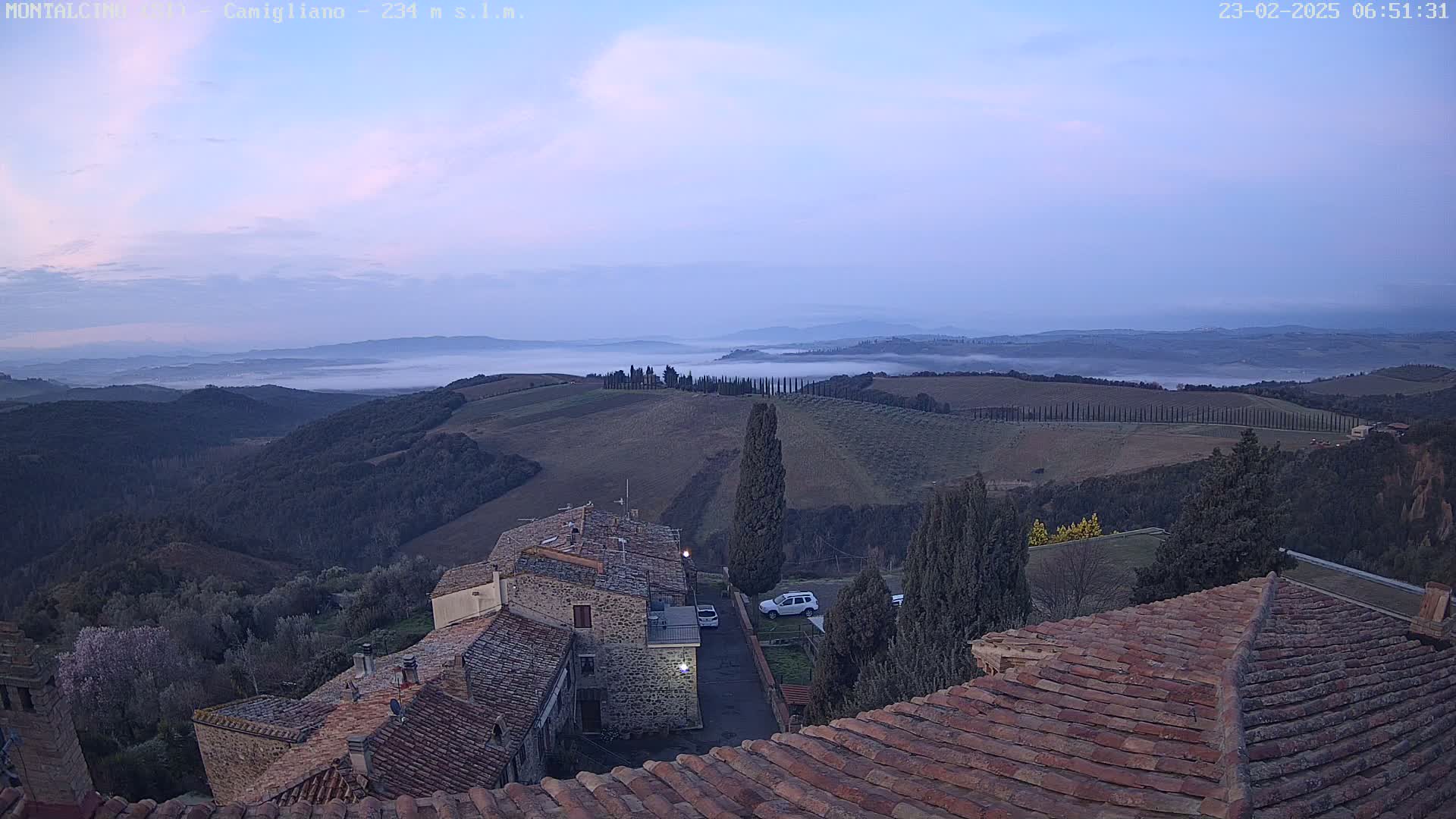 A low-lying fog blankets a valley of rolling hills and cypress trees at dawn, as seen from above a cluster of stone buildings with terracotta roofs.