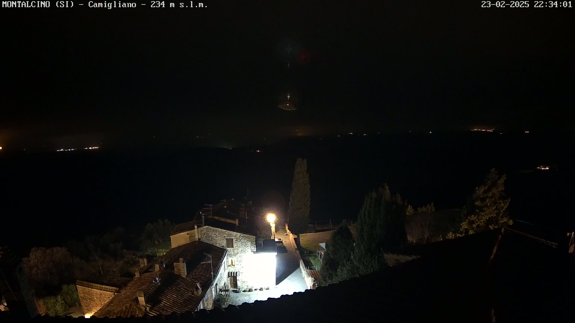 A nighttime view of a village with stone buildings illuminated by artificial light, overlooking a dark, distant landscape under a clear sky.