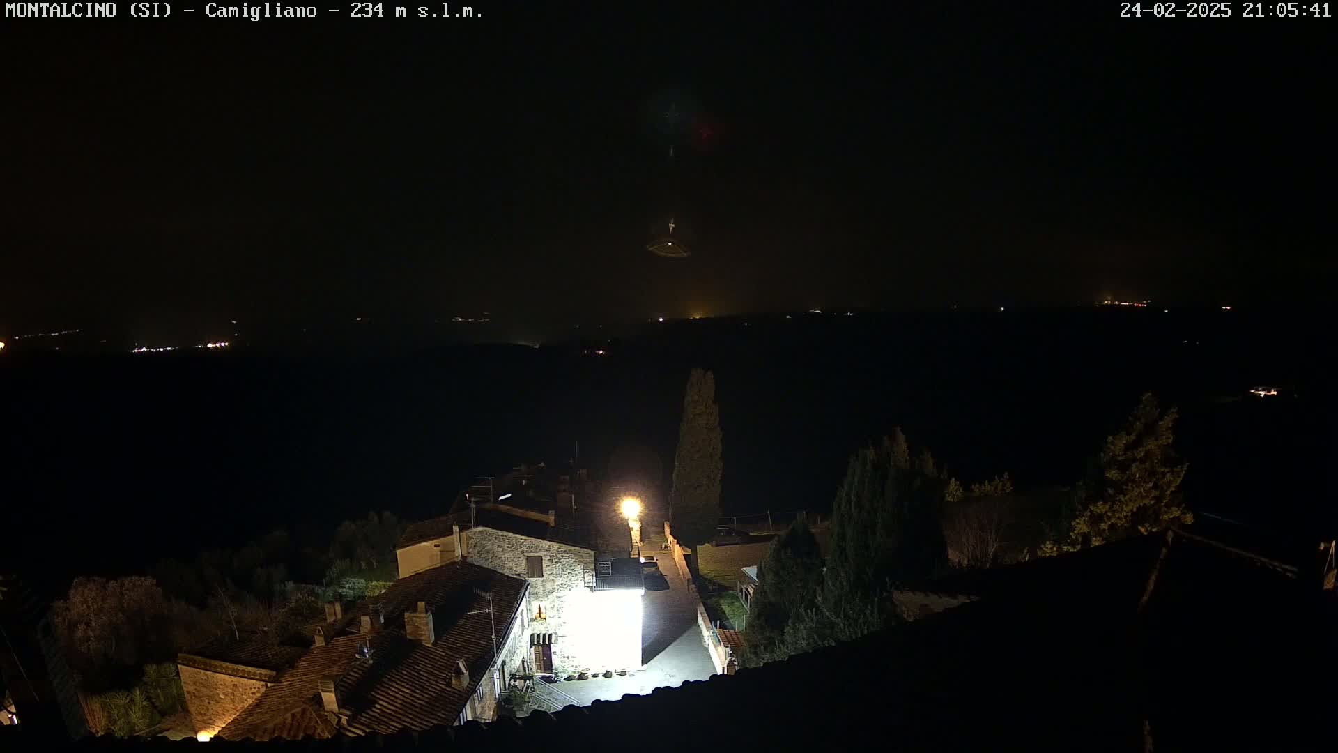 A nighttime view of a small village illuminated by a few lights, overlooking a dark, distant landscape under a clear sky.