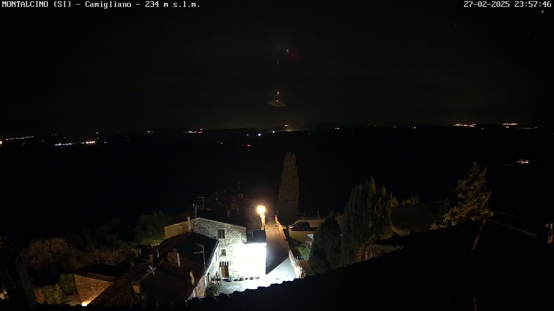 A nighttime view of a small village illuminated by lights, with a dark, clear sky showing distant lights and what appears to be a meteor.