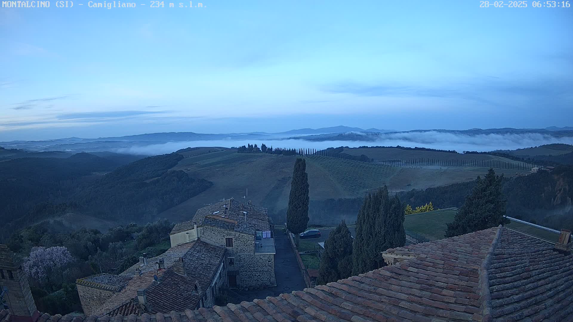 Stone buildings with terracotta roofs overlook a valley filled with fog at dawn.