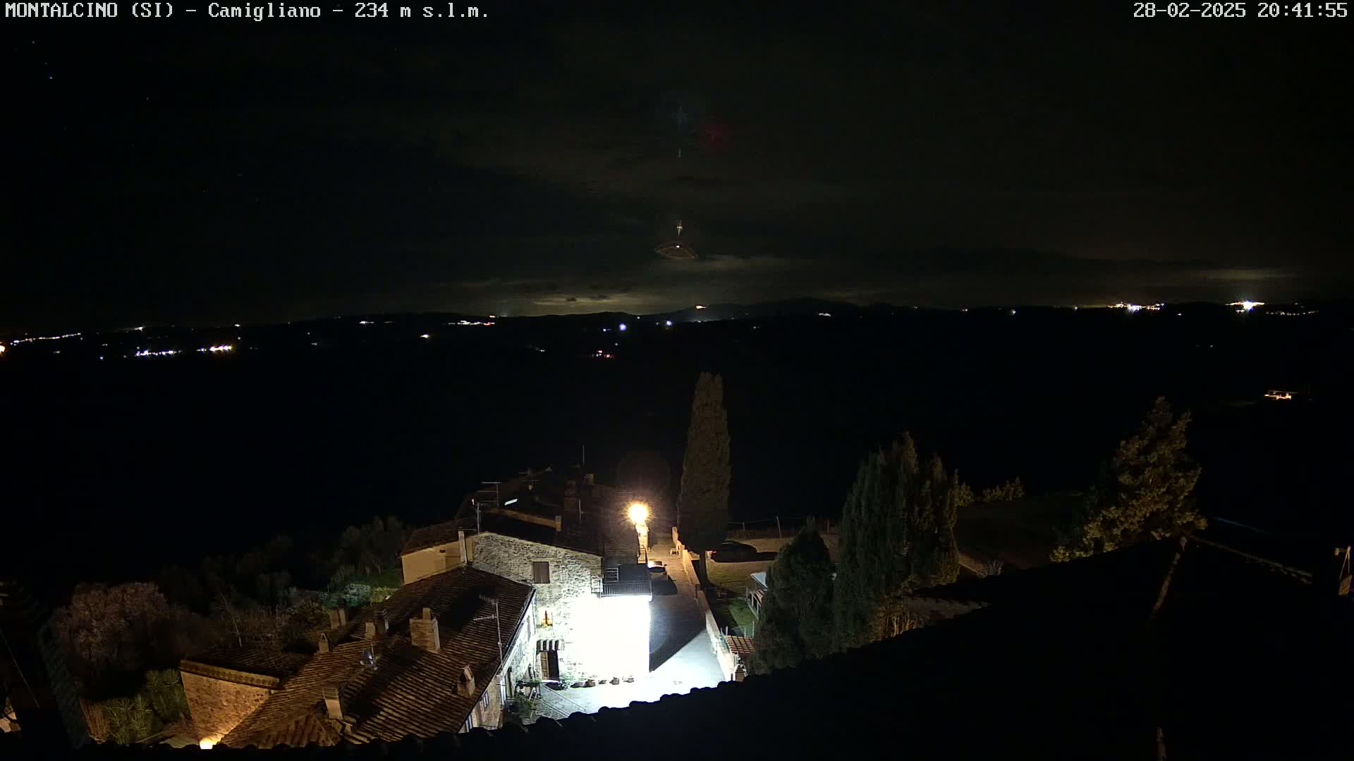 A nighttime view of a village with stone buildings, lit by streetlights, overlooking a dark valley with distant lights under a mostly cloudy sky.
