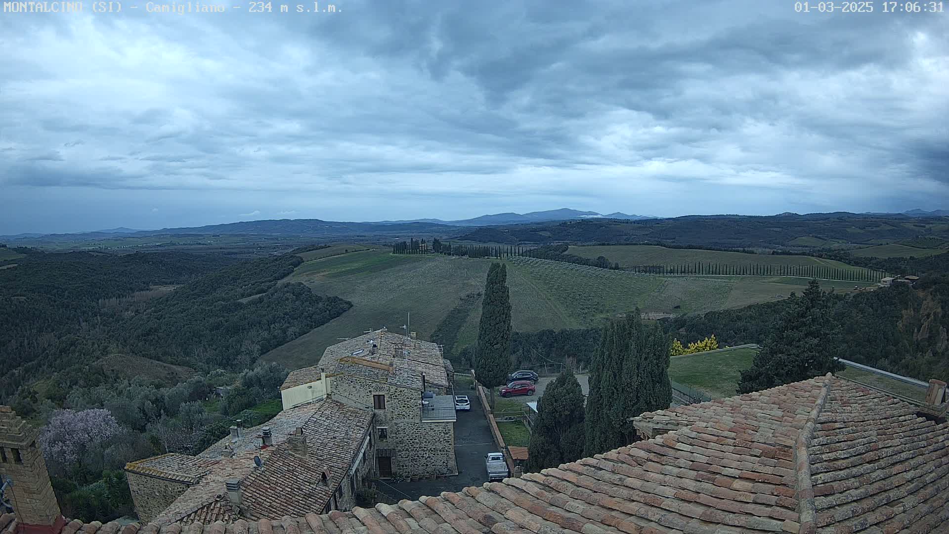 A view from a rooftop shows stone buildings, a few cars, and a sprawling landscape of rolling hills under an overcast sky.