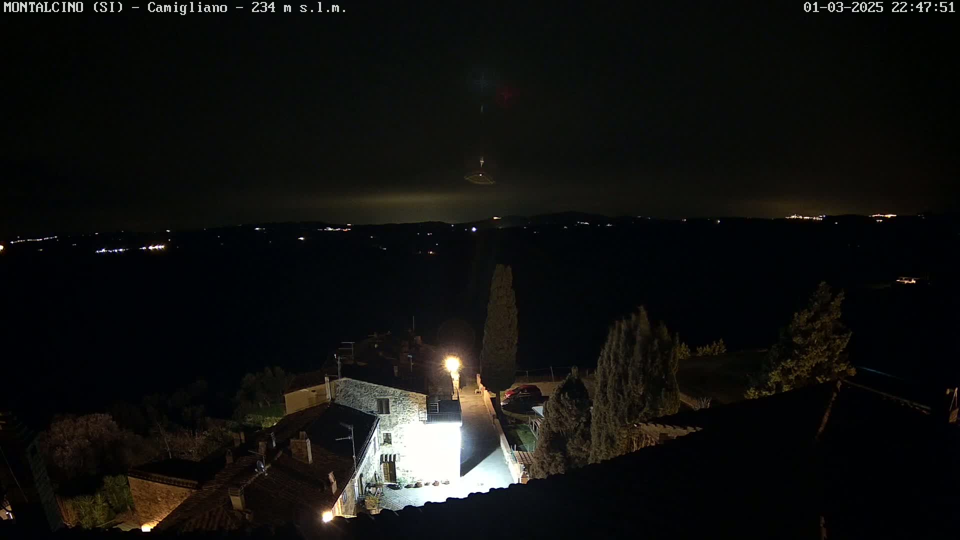A nighttime view from a rooftop shows a village with dimly lit buildings and a distant, faintly illuminated landscape under a dark sky with a possible meteor or other atmospheric phenomenon.