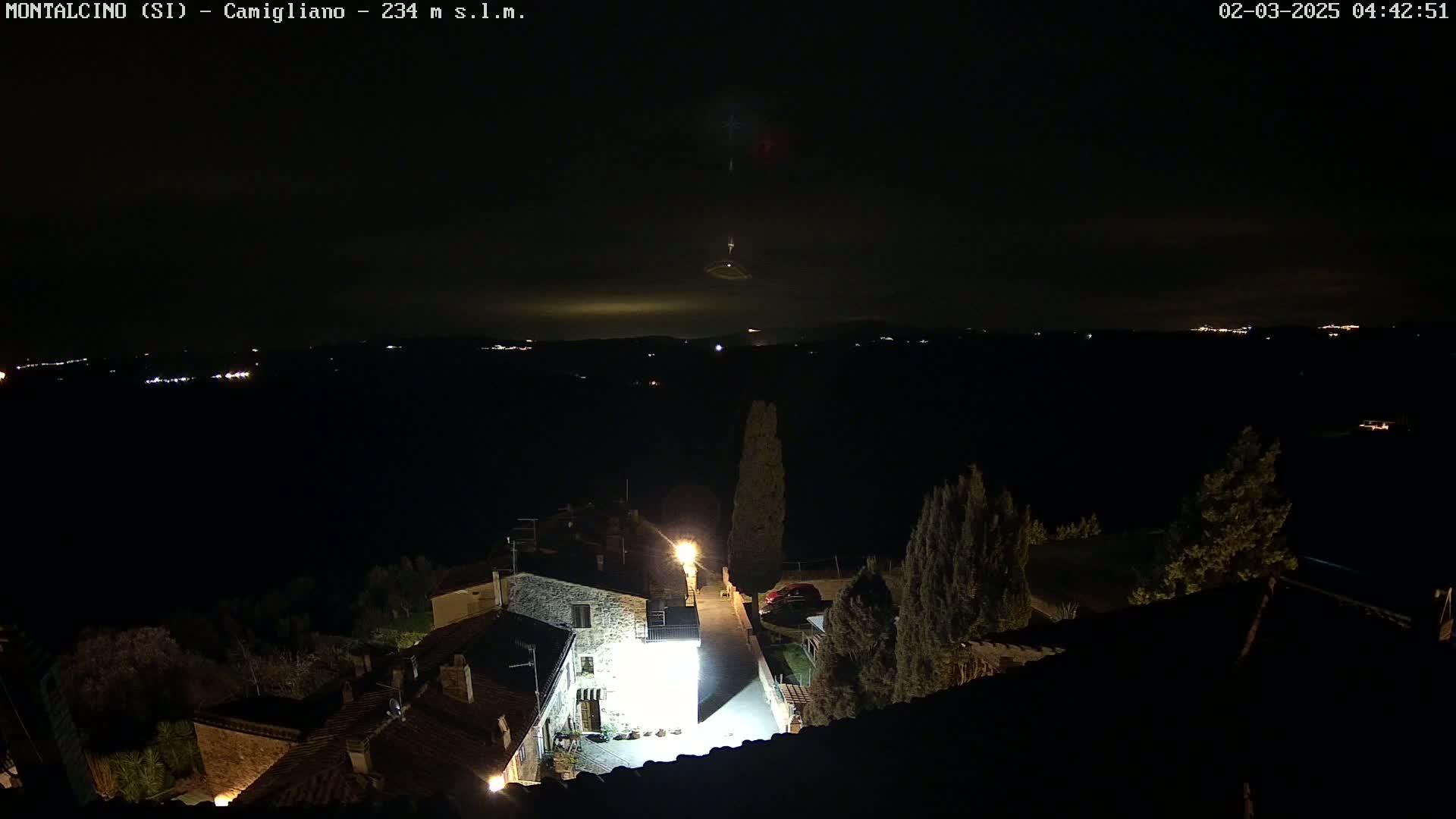 A nighttime view of a small village with dimly lit buildings, a few trees, and distant lights on a hill under a mostly clear, dark sky.