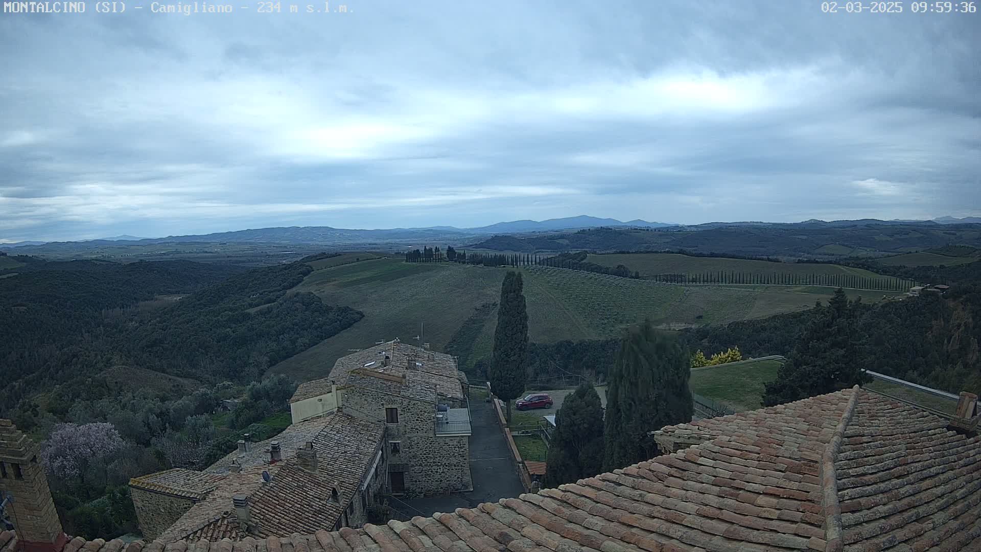 A view from atop a building with terracotta roofs shows a sprawling landscape of rolling hills, vineyards, and cypress trees under a mostly cloudy sky.