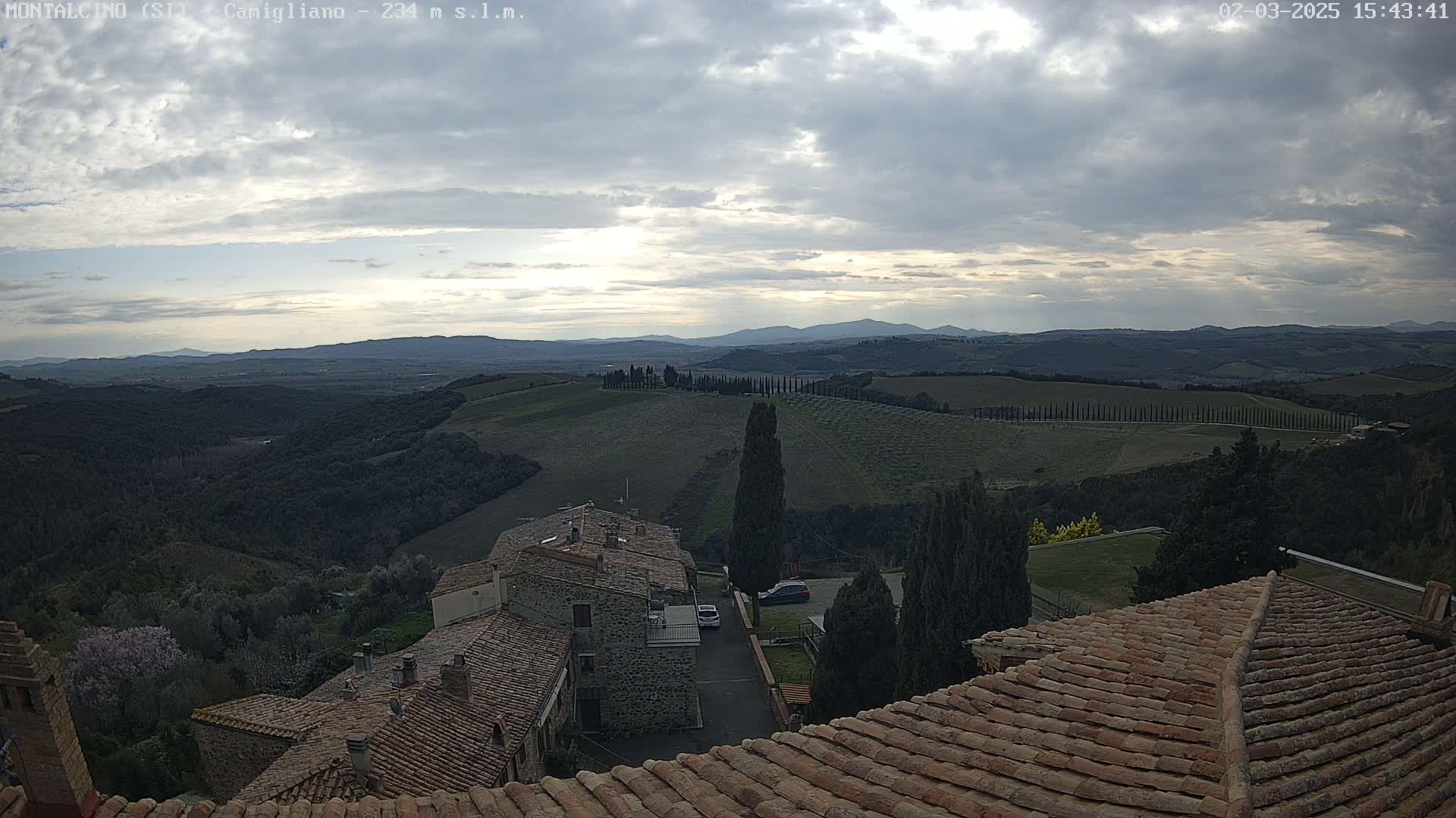 A view from a tiled roof shows a partly cloudy sky over rolling hills, trees, and buildings in a town.