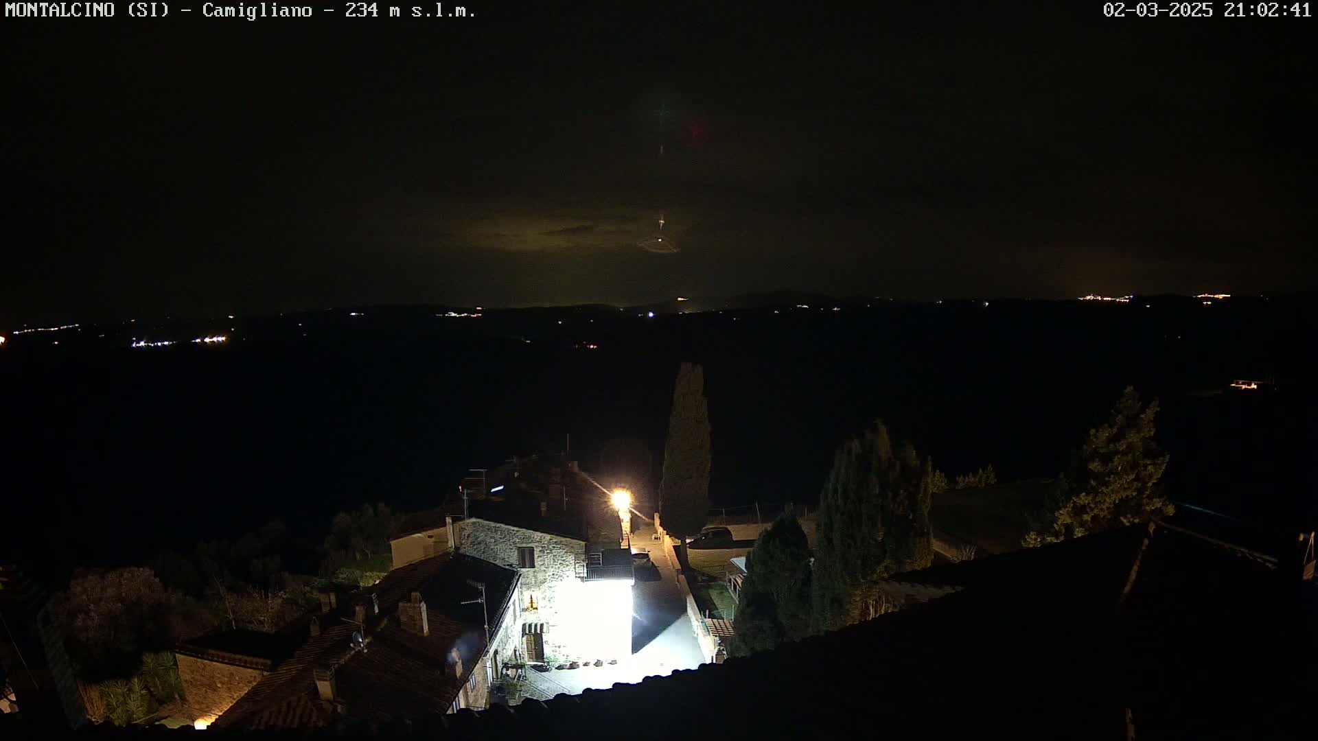 A nighttime view of a small village illuminated by streetlights, overlooking a dark landscape under a partly cloudy sky.