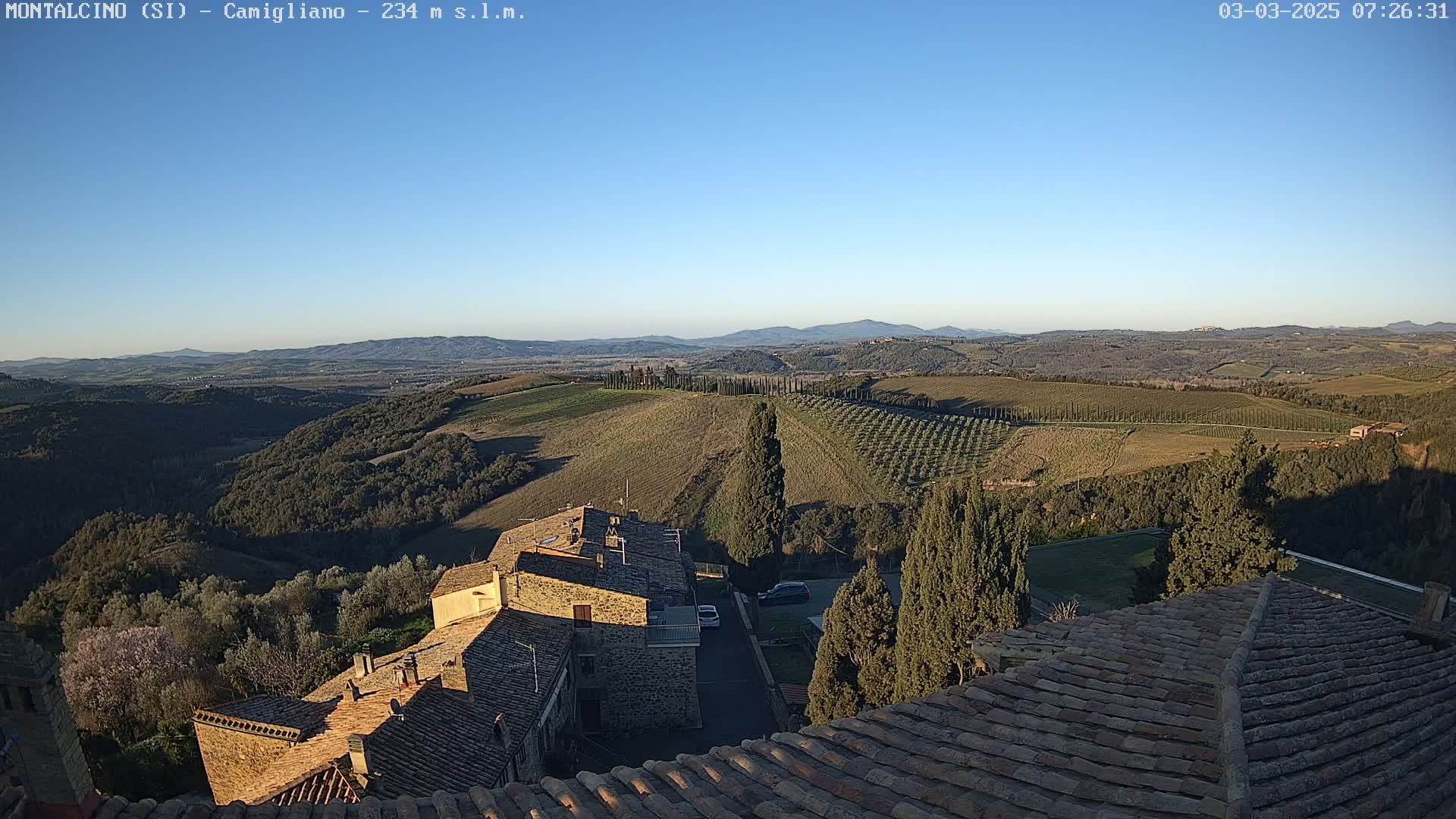 A clear, sunny day reveals a Tuscan landscape of rolling hills dotted with olive groves and cypress trees, viewed from atop a village with terracotta roofs.