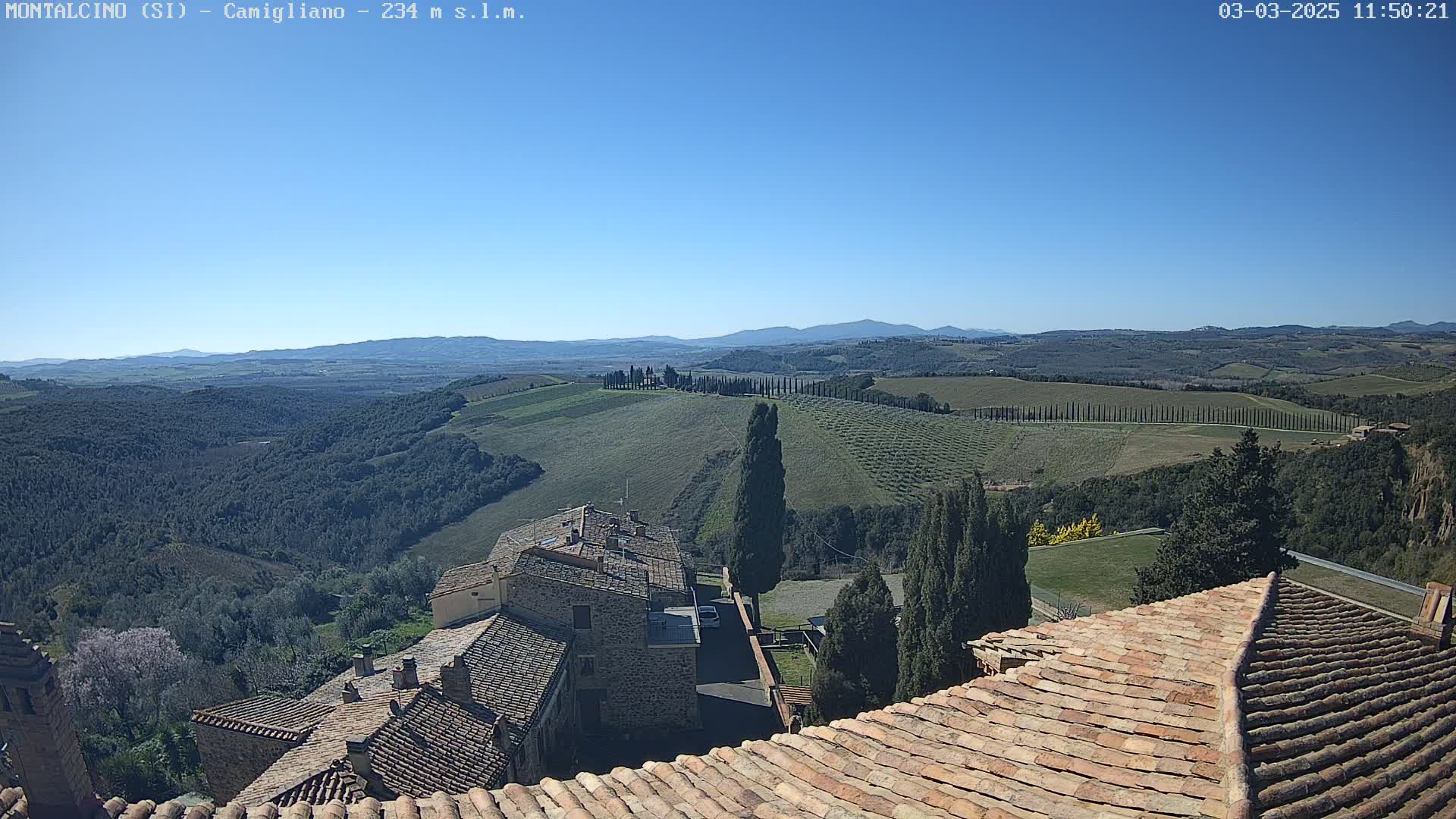 A clear, sunny day reveals a Tuscan landscape of rolling hills, cypress trees, and terracotta-roofed buildings, viewed from a high vantage point.