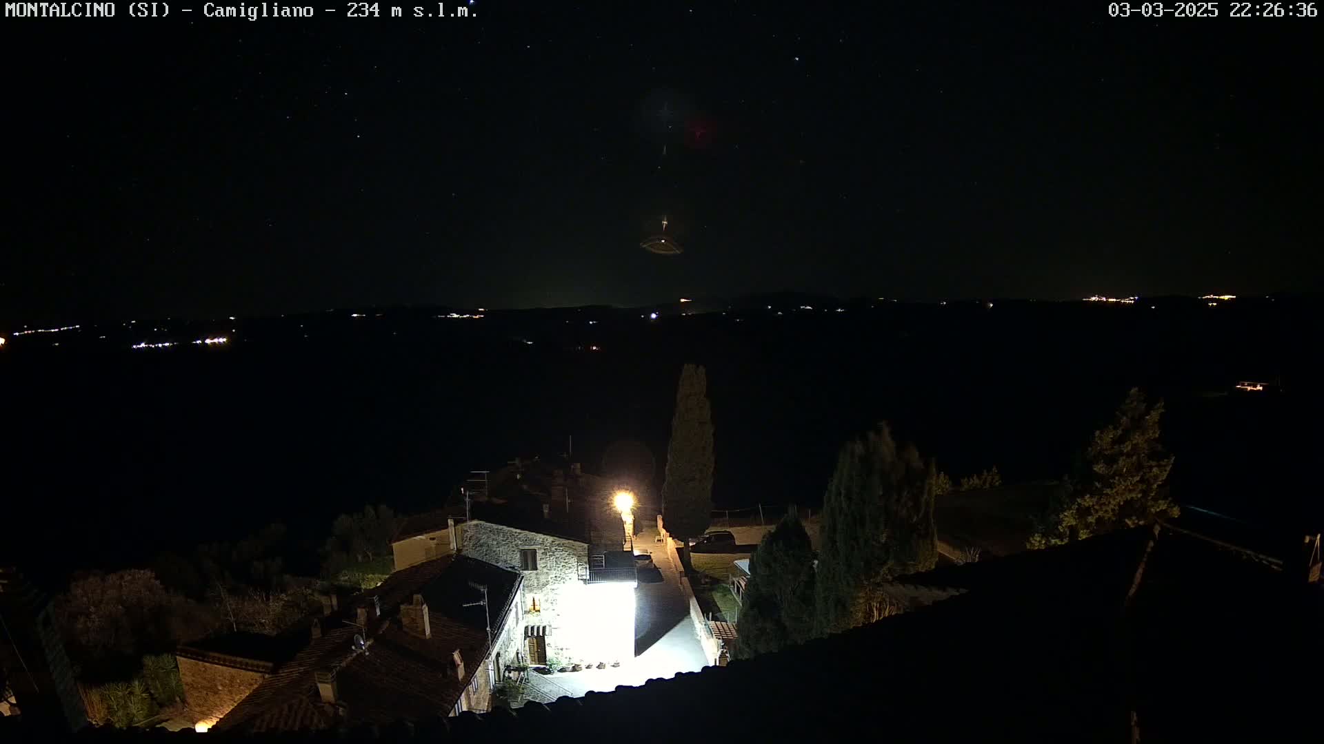 A nighttime view of a small village illuminated by streetlights, with a starry sky and distant lights visible in the background.