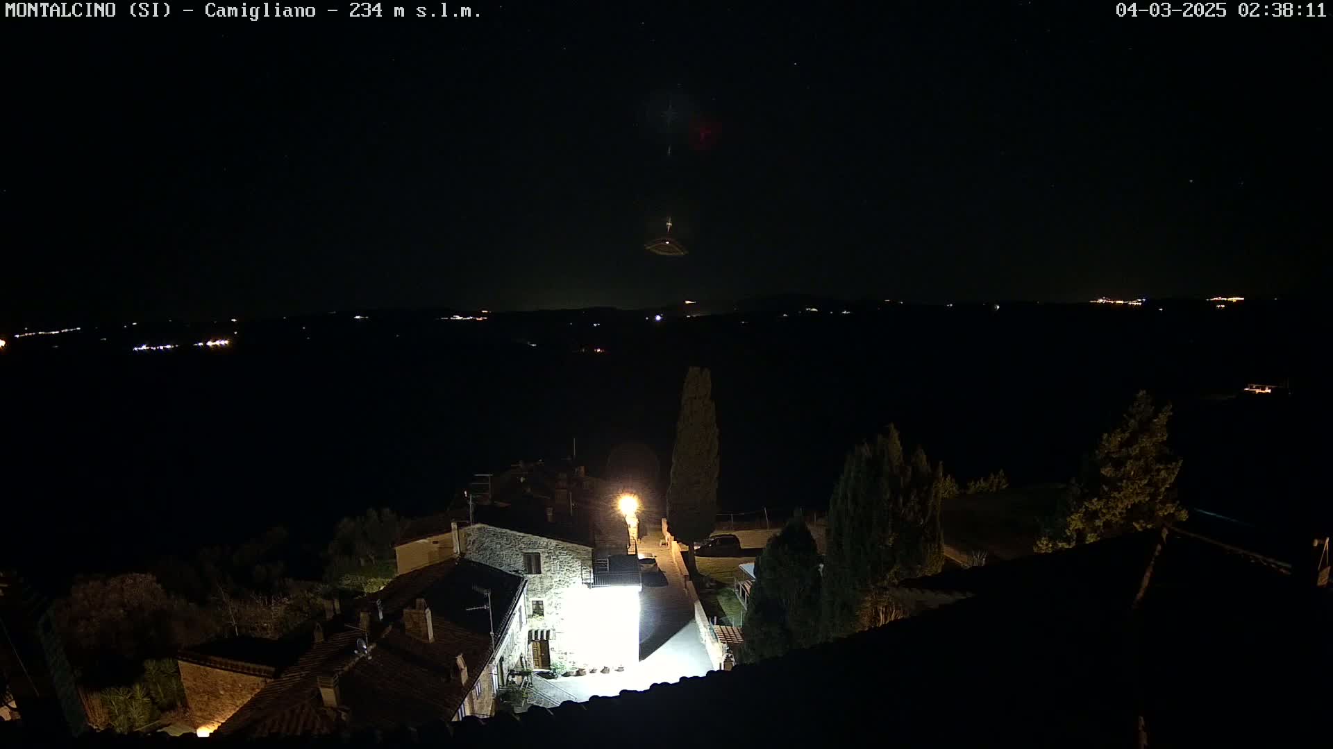 A nighttime, aerial view of a small village with a few lights on, illuminated by a bright light source, overlooking a dark landscape under a clear, starry sky.