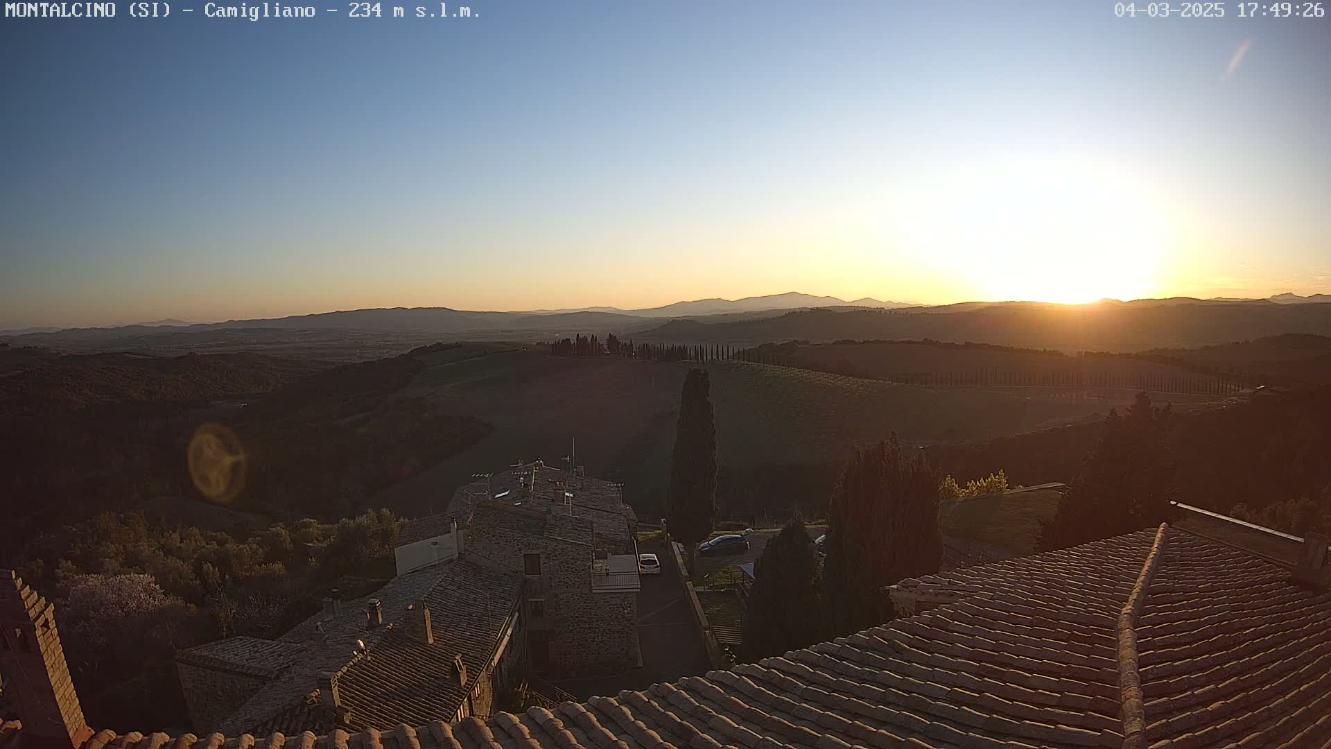 A sunset over rolling hills and a village with terracotta rooftops, under a clear sky.
