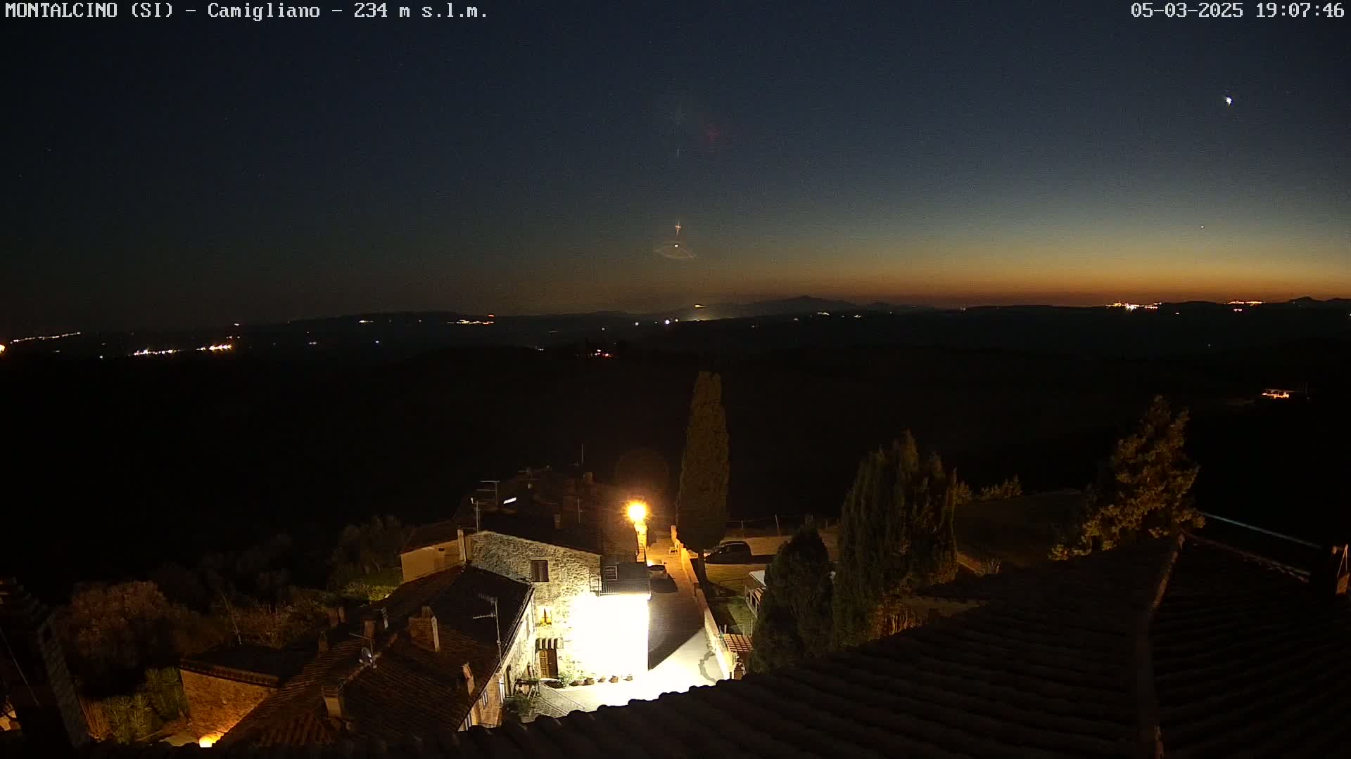 A nighttime view shows a small village nestled in a dark landscape under a twilight sky, with distant lights visible on the horizon.