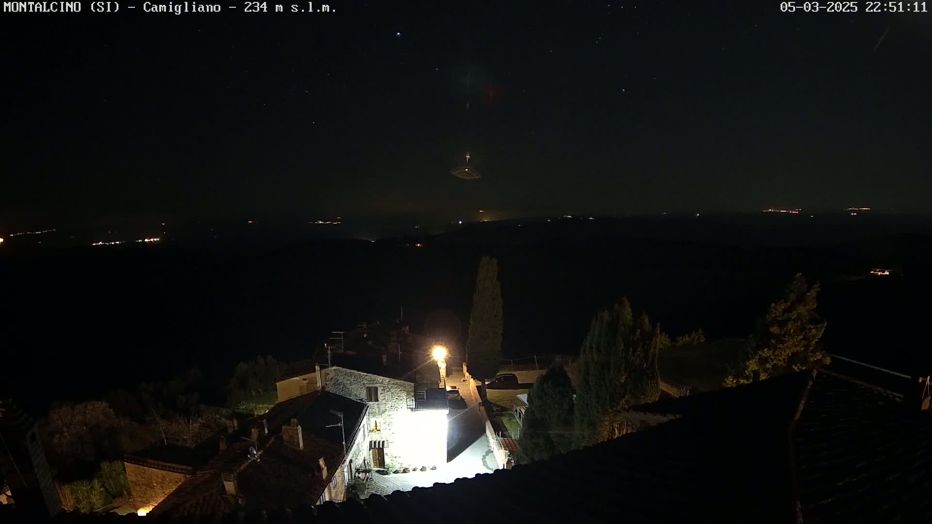 A nighttime view of a small village illuminated by streetlights, set against a dark, starlit sky with distant lights visible on the horizon.