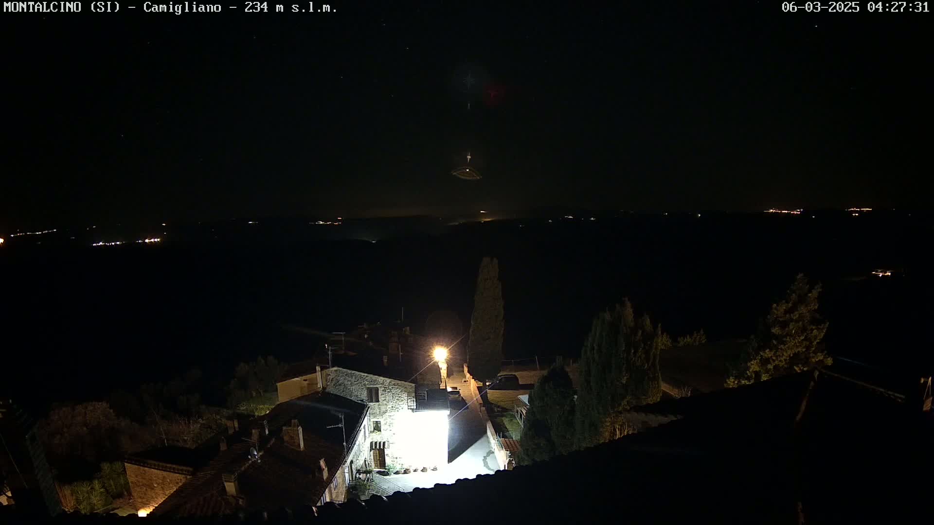 A nighttime view of a village illuminated by a few lights, with a dark, clear sky showing distant lights in the background.