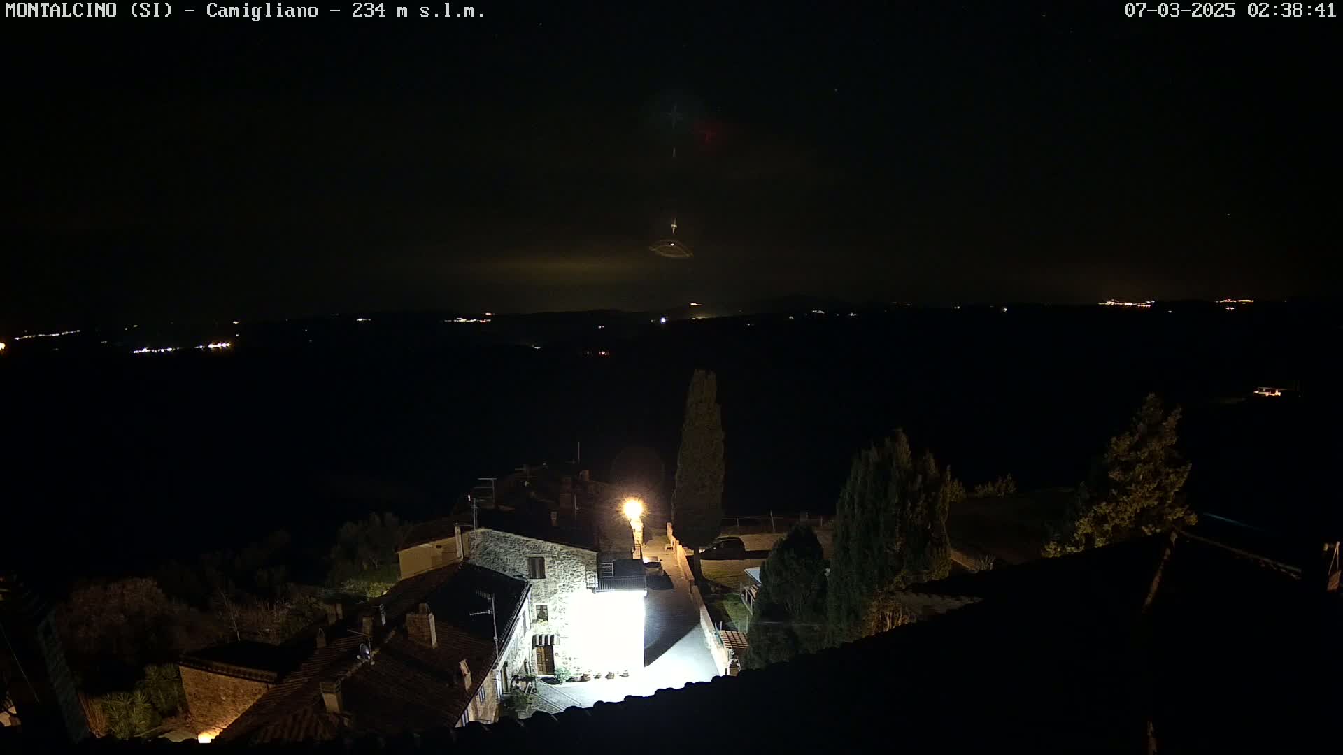 A nighttime view of a small village illuminated by streetlights, overlooking a dark, distant landscape with faint lights in the background under a mostly clear night sky.
