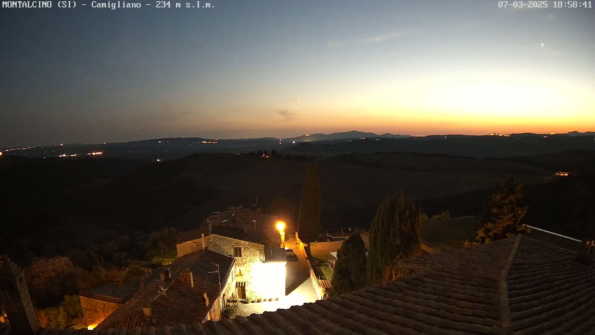 A nighttime view of a small village nestled in rolling hills, illuminated by streetlights, with a sunset visible in the distance.
