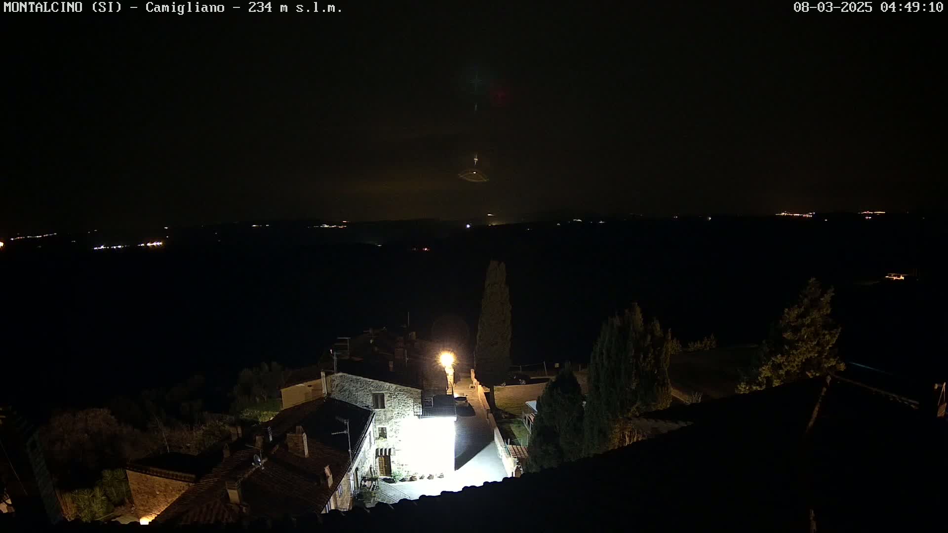 A nighttime, high-angle view of a small village illuminated by streetlights, with a distant, faintly lit horizon under a dark, possibly cloudy sky.